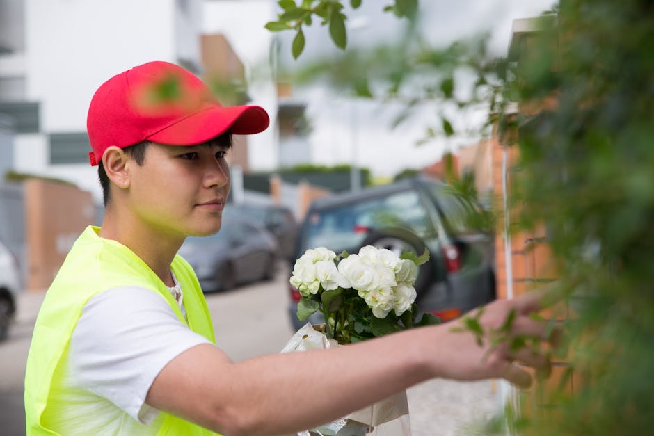 A florist wearing a red cap and neon yellow vest is delivering a floral gift to a customer on Feltham High Street. The bouquet features fresh, white hydrangeas with lush green leaves, carefully arrang
