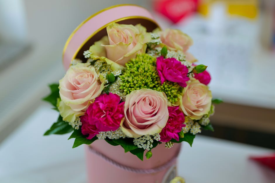 A round bouquet arrangement of fresh, pale pink roses with tightly rolled petals, complemented by vibrant magenta carnations, small white filler flowers, and bright green foliage, all inside a soft pi