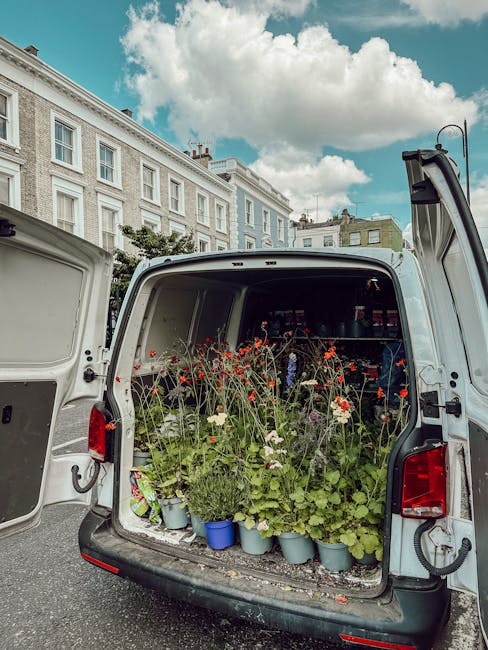The open rear of a white delivery van filled with an assortment of fresh flowering plants and bouquets, including red poppies, white daisies, and green foliage in small blue and grey pots. The arrange