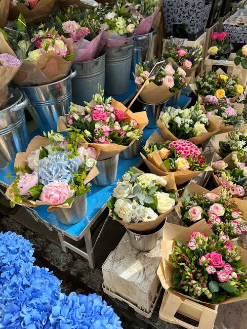 A vibrant display of fresh floral arrangements arranged in silver metal buckets and small paper-wrapped bouquets, featuring a variety of flowers including pink roses, white roses, light blue hydrangea