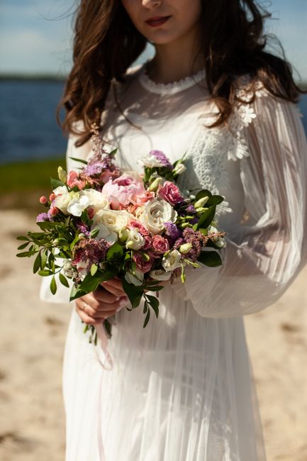 A woman in a semi-sheer, long-sleeved white dress with delicate lace accents on the collar, stands outdoors on a sandy surface near water under a partly cloudy sky. She holds a large, lush bouquet fea