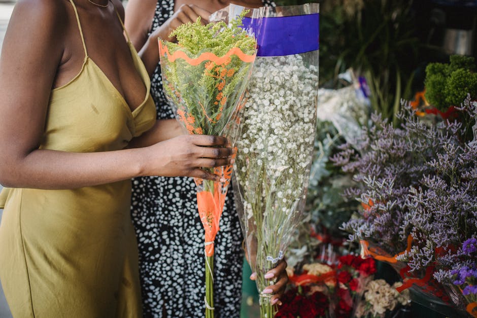 A woman in a yellow satin dress is holding a tall, cone-shaped bouquet featuring green and pale purple flowers, wrapped in clear cellophane with orange accents, in a flowershop setting. Behind her, an